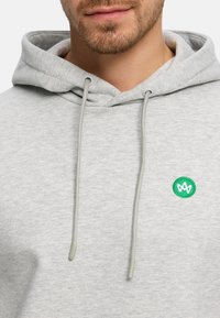 Ljusgrå huvtröja gjord av mjukt tyg. Har en känguruficka, dragsnören och en liten grön broderad logga på vänster bröst.