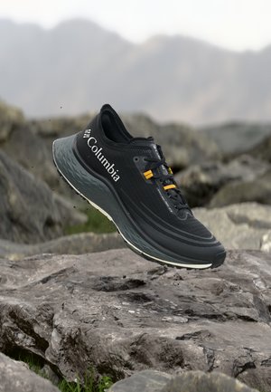 Chaussure de trail Columbia noire avec des accents jaunes sur les lacets, flottant au-dessus d'un terrain rocheux avec des montagnes floues en arrière-plan.