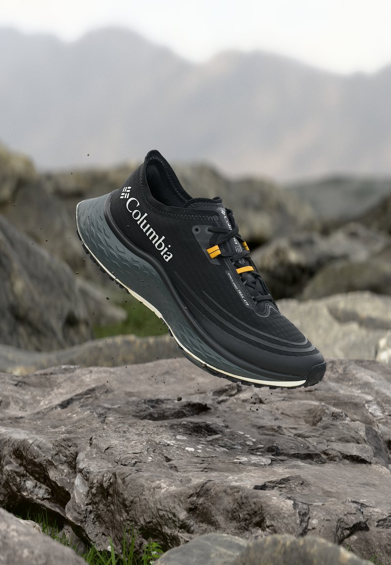 Chaussure de trail Columbia noire avec des accents jaunes sur les lacets, flottant au-dessus d'un terrain rocheux avec des montagnes floues en arrière-plan.