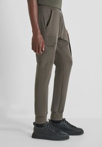 Olivgrüne Jogginghosen mit zwei seitlichen Taschen, elastischem Bund, slim fit und tapered Bein Design, kombiniert mit schwarzen Sneakers.