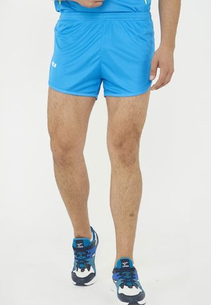Uomo con pantaloncini sportivi blu e scarpe da corsa blu, nere e bianche in piedi su uno sfondo bianco.