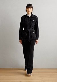 Giacca di jeans nera con vita regolabile, dotata di due tasche sul petto e bottoni argentati, abbinata a pantaloni neri a gamba larga e sneakers nere.