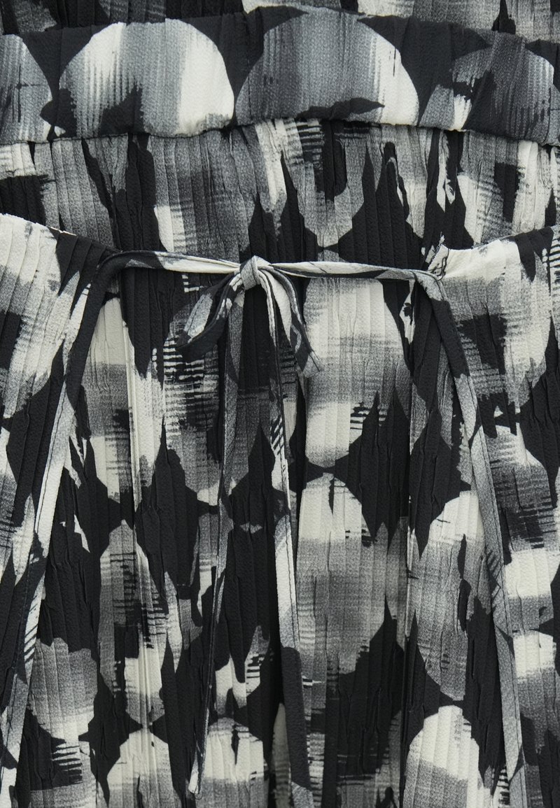 Sort-hvid plisseret stofkjole med et abstrakt mønster, med en stofsløjfe i taljen og teksturerede overfladedetaljer.