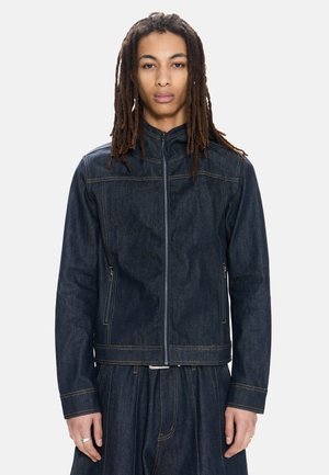 Junge Person mit langen Dreadlocks, die eine dunkelblaue Jeansjacke und passende weit geschnittene Hosen trägt, vor einem weißen Hintergrund stehend.