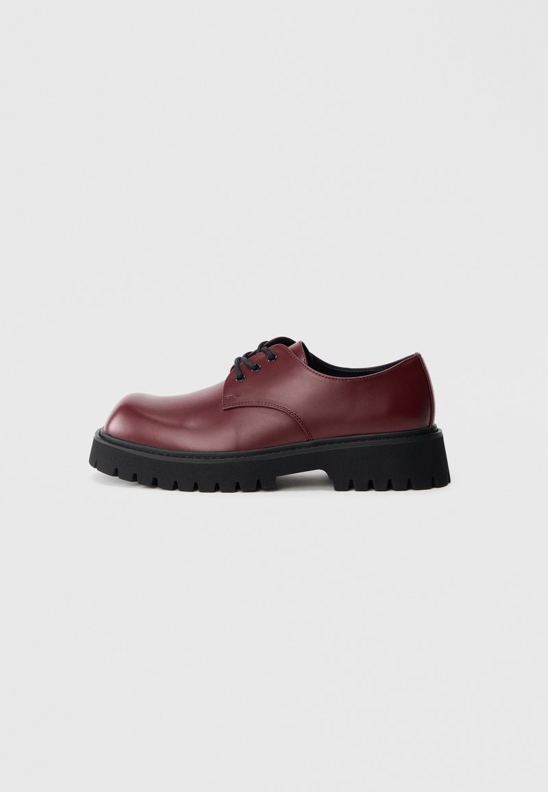 Une seule chaussure derby en cuir rouge foncé avec semelle épaisse noire à crampons et lacets noirs sur fond gris clair uni.