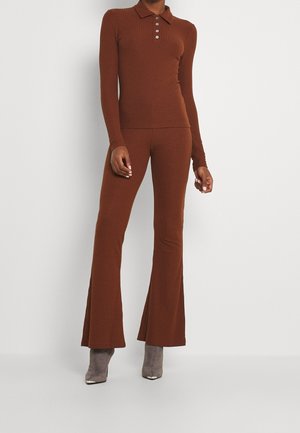 Persoon draagt een bruine geribbelde lange mouwen poloshirt en bijpassende uitlopende broek, gecombineerd met grijze suède enkellaarzen.