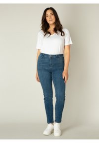 Blauwe denim skinny jeans met een middelhoge taille en een knoopsluiting, gecombineerd met een fitted witte T-shirt met korte mouwen en een V-hals, en witte sneakers.