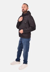 Zwarte hoodie met ritssluiting, twee voorzakken en logo op de mouw, gecombineerd met blauwe jeans en witte sneakers.