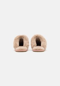 Suddiga beige tofflor med öppen baksida, mjukt pälsliknande material, platt rosa sula och rundad form, lämpliga för inomhusbruk.