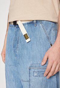 Svetlo modri denim hlače z ohlapnim krojem, ki imajo teksturirano površino, kremni pas in vidne šive.