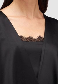 Vêtement en satin noir avec un décolleté en V profond, superposé à un haut orné d'une bordure en dentelle noire le long de l'encolure. Texture lisse sur toute la surface.