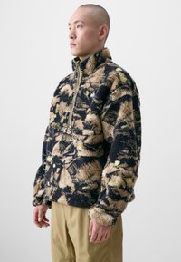 Fleece pullover with a high collar, featuring a black, beige, and green camouflage pattern. Zippered front and side pocket detail.