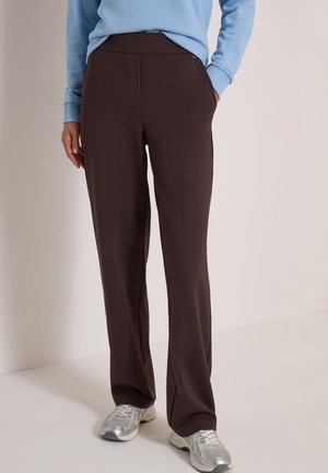 Personne portant un pantalon droit brun foncé, un pull bleu clair et des chaussures de sport argentées, debout avec une main dans la poche contre un mur uni.