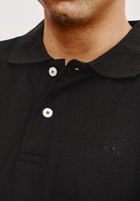 Czarna koszulka polo z kołnierzykiem, z trzema białymi guzikami i małym logo po lewej stronie. Wykonana z teksturowanej tkaniny.