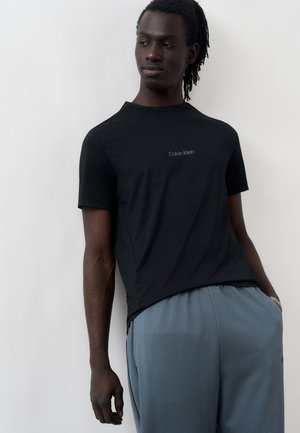 Homme à la peau foncée et aux dreadlocks, portant un t-shirt noir Calvin Klein et un pantalon gris, debout contre un mur blanc, les mains dans les poches.