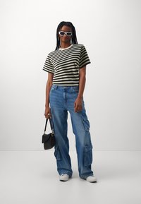 T-shirt court à rayures noires et blanches associé à un jean cargo bleu ample avec des poches, des baskets blanches et un petit sac à main noir.