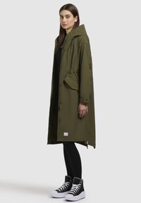 Parka longue verte olive avec une capuche, taille réglable par cordon, poches latérales, fermeture à boutons-pression, assortie à des leggings noirs et des baskets montantes.