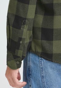 Camicia di flanella a quadri verdi e neri con polsini abbottonati, texture morbida e orlo curvato verso l'alto. Indossata con jeans in denim blu.