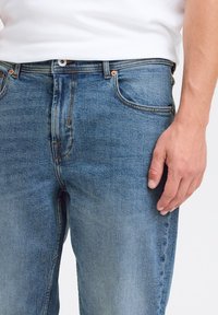 Blå denimjeans med rette ben, synlige sømmer og kobberdetaljer, med en hånd som hviler på linningen.