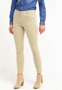 Pantalons skinny beige avec une texture lisse, poches avant et taille mi-haute, associés à des bottes de cheville beige clair avec un talon en bois.