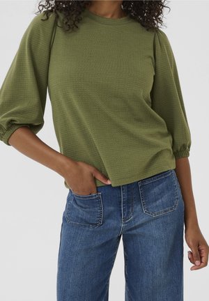 Femme portant un chemisier texturé vert olive à manches bouffantes trois-quarts et un jean en denim bleu avec des poches plaquées à l'avant.