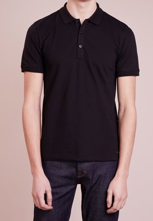 Man wearing a plain black short-sleeve polo shirt and dark denim jeans, standing against a neutral background.
