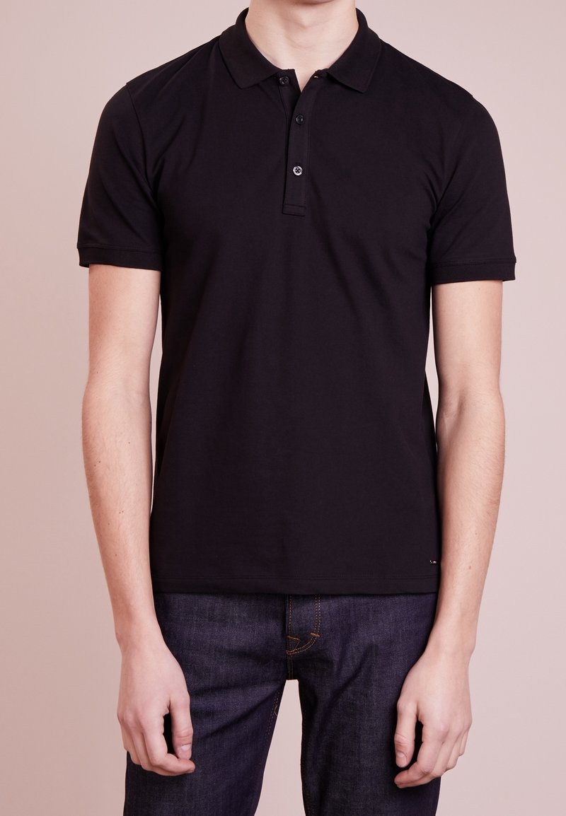 Homme portant un polo noir uni à manches courtes et un jean en denim foncé, debout devant un fond neutre.