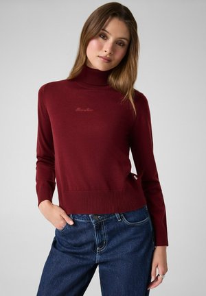 Junge Frau trägt einen burgunderroten Rollkragenpullover und blaue Jeans, posiert mit einer Hand in der Tasche vor einem einfarbigen grauen Hintergrund.