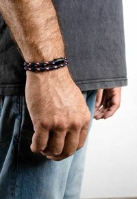 Tkaný náramok v čiernej, červenej a bielej farbe s textúrovaným povrchom. Obsahuje čiernu pracku, nosený na zápästí. Viditeľný predlaktie a košeľa.