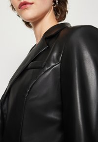 Musta nahkablazer, jossa on terävä kaulus, rakenteelliset olkapäät ja hienovarainen kiilto. Tarkkaompeleet ja kapea istuvuus korostavat mallia.