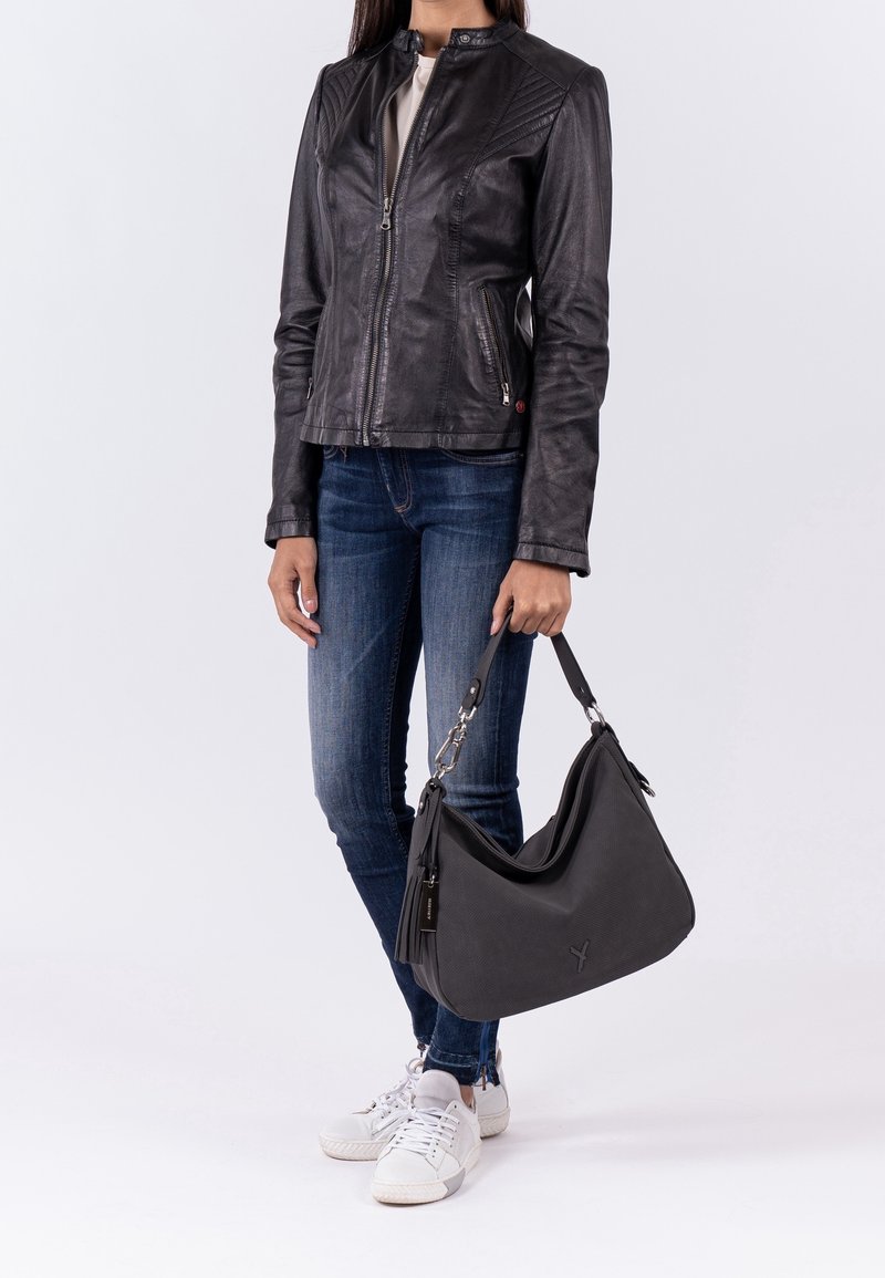Femme portant une veste en cuir noire, un jean bleu, des baskets blanches et tenant un grand sac à main noir sur un fond uni.