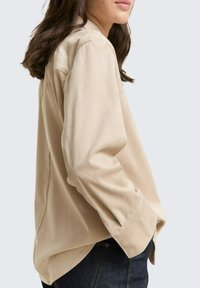 Camisa de manga larga beige con un corte holgado, que presenta una textura suave, puños abotonados y un bolsillo lateral, combinada con jeans oscuros.