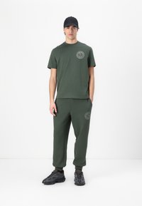 Camiseta de algodón verde oliva y pantalones jogger a juego con acentos de logo. Los joggers tienen puños elásticos. Zapatillas negras completan el conjunto.