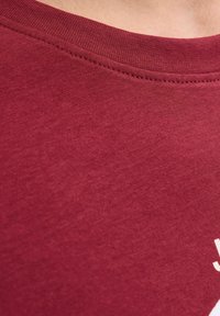 Primer plano del pecho superior de una persona que lleva una camiseta de cuello redondo en color burdeos, con costuras visibles a lo largo del escote.