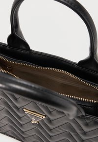 Μαύρη δερμάτινη τσάντα με καπιτονέ σχέδιο, με χρυσό λογότυπο Guess, διπλές λαβές και εμφανές ανοιχτό φερμουάρ που αποκαλύπτει το εσωτερικό με ανοιχτό καφέ φόδρα.