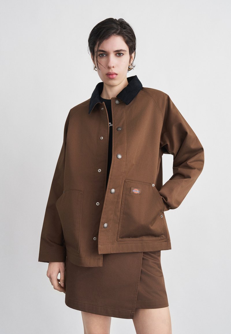 Νεαρός με κοντά σκούρα μαλλιά, φορώντας καφέ μπουφάν με κουμπιά και μαύρο γιακά, καθώς και καφέ φούστα που ταιριάζει, σε απλό φόντο.