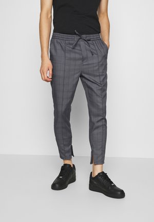 Osoba w szarych, kratkowanych spodniach z troczkami i zamkami przy kostkach oraz czarnych trampkach, stoi na tle jednolitego jasnego tła.