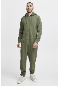 Mono de cremallera de color verde oliva en tela suave, con capucha, dos bolsillos frontales y puños acanalados. Combinado con zapatillas blancas.