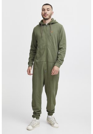 Olive green zip-up onesie in soft fabric, featuring a hood, two front pockets, and ribbed cuffs. Paired with white sneakers.