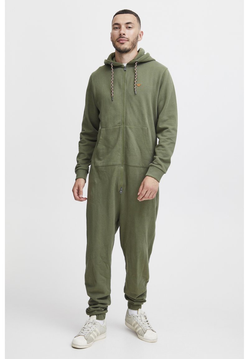 Oliven grønn zip-up heldress i mykt stoff, med hette, to frontlommer og ribbede mansjetter. Kombinert med hvite joggesko.