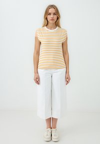 Gestreepte mouwloze top in wit en oranje, gecombineerd met brede witte broeken. Het model draagt crèmekleurige sneakers.