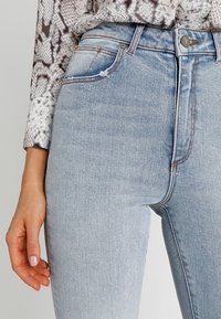 En hand med ljusrosa nagellack vilar nära fickan på ljusblå högviktsjeans, iförd en långärmad topp med orm-mönster.