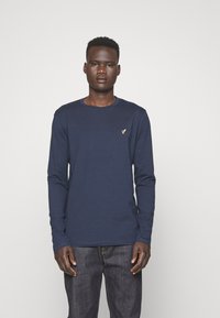 Tricou din bumbac cu mânecă lungă de culoare bleumarin, cu guler rotund și un mic detaliu cu logo auriu pe piept, asortat cu blugi denim întunecați.