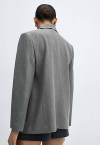Blazer gris con un diseño estructurado, que presenta un cuello con muescas, ajuste entallado y tela texturizada. Detalle del respiradero trasero visible.