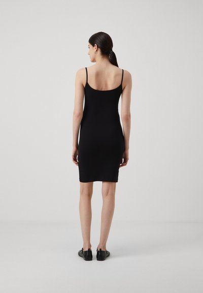 Femme de dos portant une robe noire ajustée sans manches et des chaussures plates noires, se tenant devant un fond clair et uni.