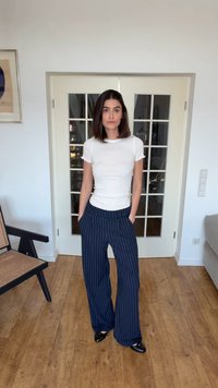 Žena stojaca v interiéri, oblečená v bielej priliehavej tričku, tmavomodrých širokých nohaviciach s prúžkami a čiernych topánkach, ruky v kapsách, neutrálna mimika.