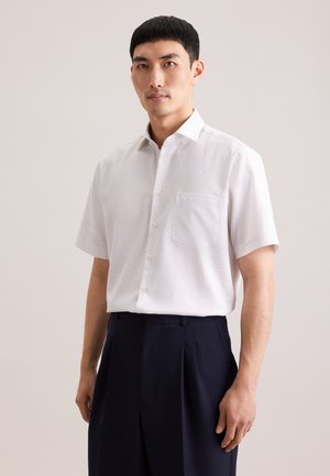 Mand iført en hvid kortærmet skjorte med småmønstret tryk og mørke, plisserede bukser, stående foran en ensfarvet baggrund.