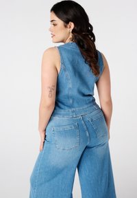 Een vrouw met golvend haar poseert in een mouwloze denim jumpsuit, waarbij ze een bloemtattoo op haar arm laat zien tegen een witte achtergrond.