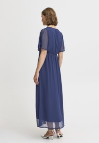 Mörkblå maxiklänning med genomskinliga ärmar, sammanknuten midja och flödande tyg. Noterbar detalj med genomskinlig nederkant. Stylerad med vita klacksandaler.