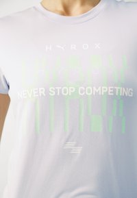Hele lilla sportline t-särk, mis on valmistatud siledast ja venivast materjalist. Omab valget ja rohelist teksti: "HYROX NEVER STOP COMPETING," koos mustritega.